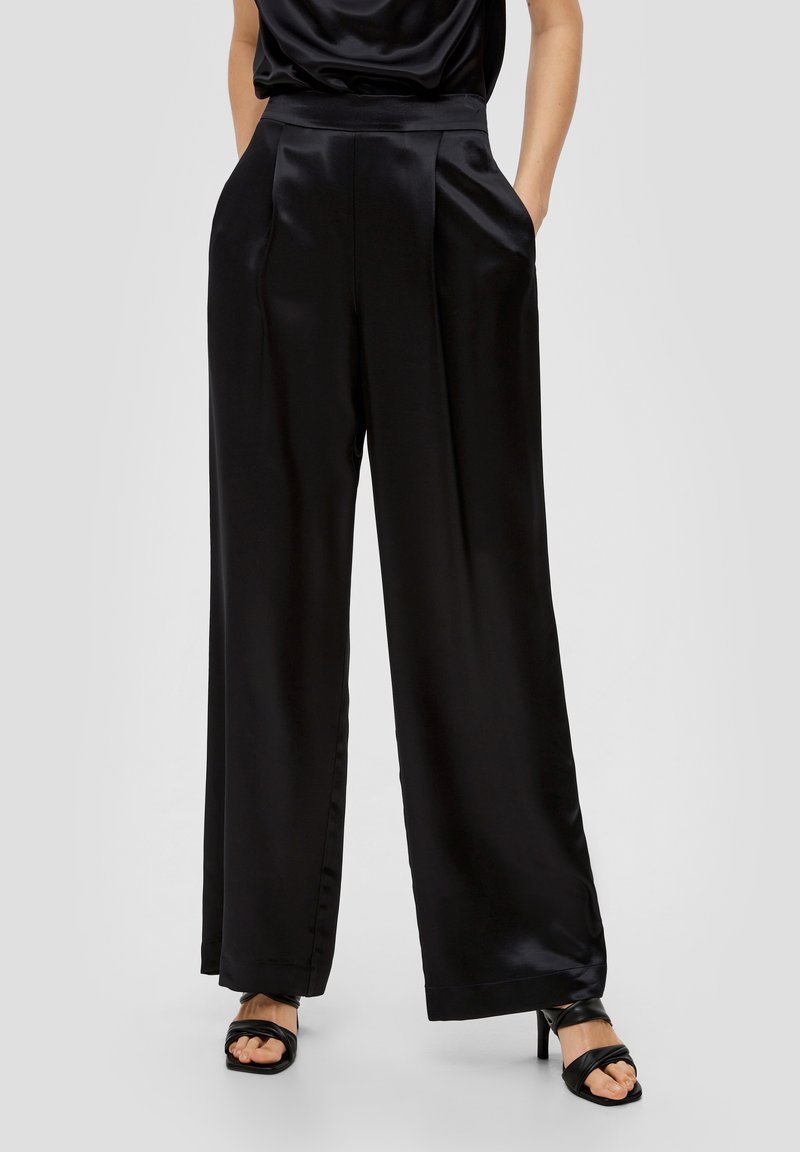 Pantalon large en satin noir avec des plis sur le devant, deux poches latérales et une texture lisse, associé à des sandales à talons strappy noires.
