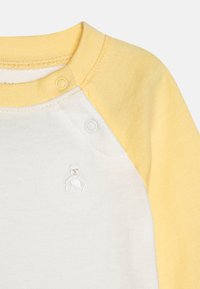 Koksfarget skjorte med gule raglan-sleeves og hvit kropp, med et lite brodert bjørne-design på brystet og trykknapper på skulderen.