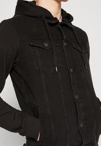 Svart denimjacka med en mjuk tyghuva, med framfickor, knappar och kontrasterande texturer. Närbild av den övre halvan.