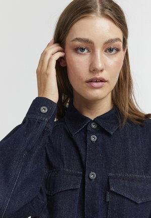 Dunkelblauses Denimhemd mit Kragen, Knopfleiste und zwei Brusttaschen. Mit Metallknöpfen, strukturiertem Stoff und lockerer Passform.