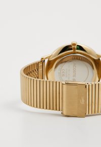 Zelta metāla pulkstenis ar teksturētu siksniņu, piedāvājot apaļu korpusu un sprādzi. Aizmugurē ir redzams zīmola logo un modeļa informācija.
