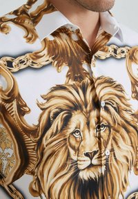 Homme portant une chemise blanche ornée de motifs détaillés de lion doré et marron et de chaînes ornées, montrant le col et une partie du visage.