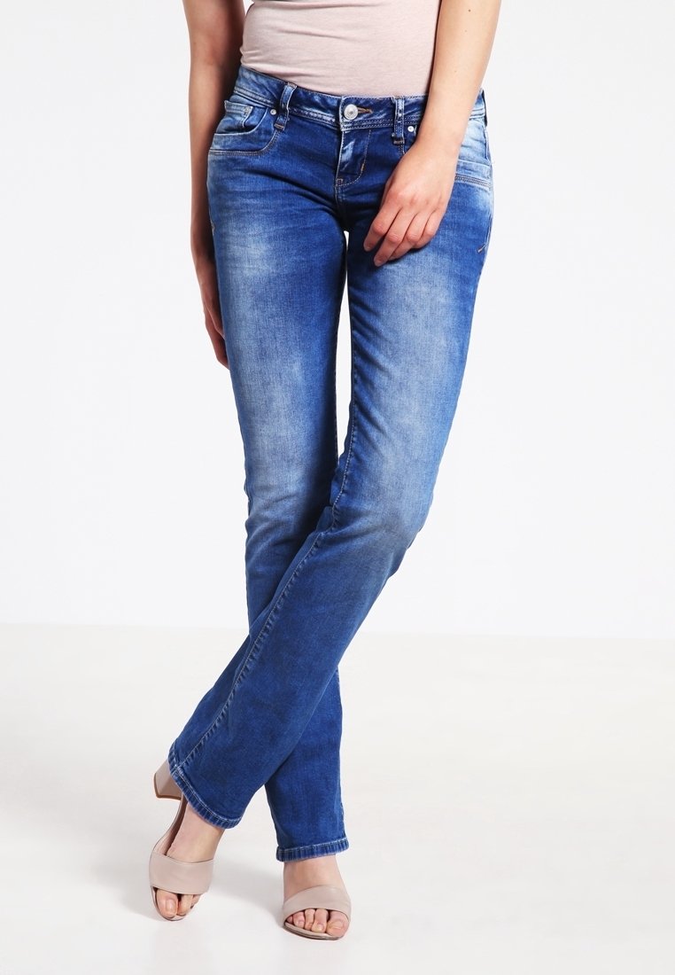 Jeans en denim bleu avec un effet délavé, ajustés au niveau des cuisses et légèrement évasés à l'ourlet, associés à des sandales à talons de couleur claire.