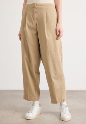 Personne portant un pantalon beige à larges jambes, des chaussettes blanches et des baskets blanches, debout sur un sol gris devant un fond uni.