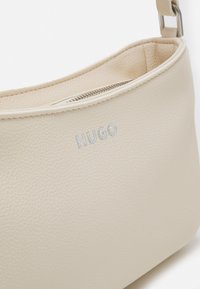 Beige leather handbag with a structured shape, featuring a zip closure and embossed "HUGO" logo in silver. Smooth texture with subtle grain.