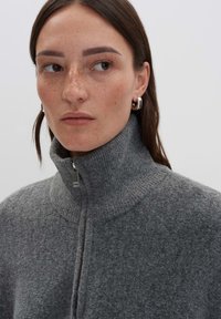 Frau mit braunen Haaren und Sommersprossen, die silberne Creolen und einen grauen Wollpullover mit Reißverschluss und hohem Kragen trägt, schaut zur Seite.