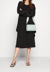 Černý dlouhý kardigan přes černý top a kalhoty s širokými nohavicemi, doplněné černými žabkami. Světle modrá kabelka přes rameno dodává kontrast.