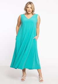 Ein türkisfarbenes, ärmelloses Maxikleid mit V-Ausschnitt, ausgestattet mit Seitentaschen und einer fließenden Silhouette, aus weichem, leichtem Stoff gefertigt.