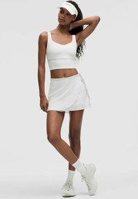 Top corto blanco con tirantes anchos, combinado con una falda envolvente blanca que presenta un lazo lateral. La modelo lleva una visera blanca y zapatillas deportivas blancas.