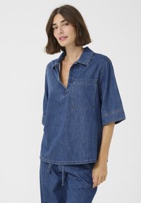 Denim bluza s sproščenim krojem, ovratnikom, kratkimi rokavi in žepom na prsih. Dopolnjena z vidnimi šivi v kontrastnih barvah.