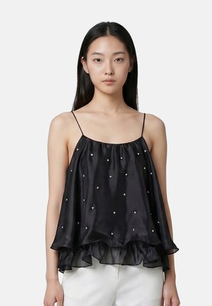 Giovane donna con lunghi capelli neri che indossa una canotta nera larga senza maniche con piccoli ornamenti bianchi e pantaloni bianchi.