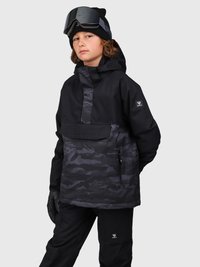 Joven vestido con una chaqueta de invierno de camuflaje negro y gris con capucha, pantalones de nieve negros, guantes, un gorro y gafas de esquí en la cabeza.
