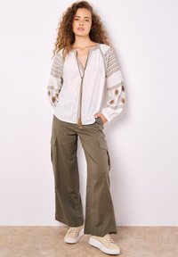 Weiße Bluse mit goldener Stickerei und Quasten-Detail; kombiniert mit olivgrünen Cargo-Hosen; Modell trägt beige Plateausneaker.
