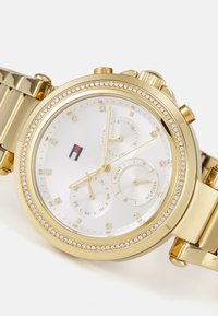 Orologio placcato oro con quadrante bianco, lunetta decorata con cristalli, tre sottoquarti e bracciale in acciaio inossidabile. Logo visibile sul quadrante.