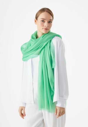 Écharpe verte transparente en tissu léger, drapée de manière décontractée, avec une texture douce et des bords frangés, portée sur un top blanc.
