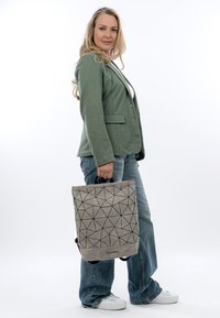 Mujer con cabello largo y rubio, vestida con blazer verde, jeans azules y zapatillas blancas, sosteniendo una mochila gris con patrón geométrico por el asa.