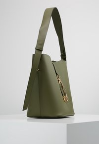 Olivově zelená kožená kabela ve tvaru kbelíku s širokým nastavitelným ramenním popruhem a zlatým řetízkovým detailem na přední straně, vystavená na bílé podstavci.
