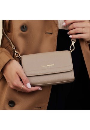 Beige leather crossbody bag with gold logo, rectangular shape, flap closure, and adjustable strap, held against a brown coat backdrop.
