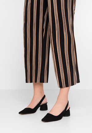 Černé semišové slingback boty s špičatou špičkou a mírným blokovým podpatkem, spárované s černými a oranžovými pruhovanými zkrácenými kalhotami.