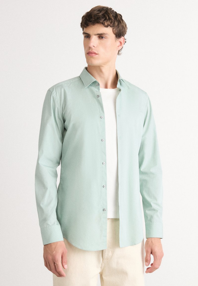 Căma�șă verde deschis cu mânecă lungă, guler cu nasturi, textură netedă, nasturi pe față și o croială lejeră, purtată peste o cămașă albă.