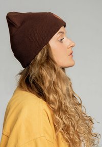 Brązowa dziana czapka typu beanie z wyraźnym, spiczastym wierzchołkiem i widoczną fakturą ribbed, z okrągłą metką z logo, noszona przez osobę z kręconymi, długimi włosami.