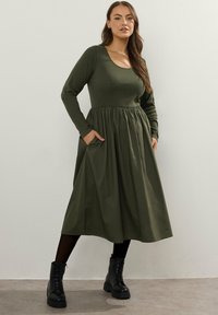 Kobieta w oliwkowej, długiej sukience midi z marszczoną talią i kieszeniami, w połączeniu z czarnymi rajstopami i butami na sznurówki.