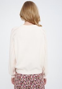 Maglione beige chiaro con texture lavorata a maglia, maniche a sbuffo e dettagli traforati sulle spalle, abbinato a una gonna con motivo floreale.
