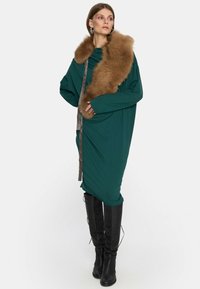 Kobieta ubrana w luźną, ciemnozieloną sukienkę z długimi rękawami, dużą brązową futrzaną stole, oraz czarnymi botkami do kolan, stojąca z jedną ręką na biodrze.