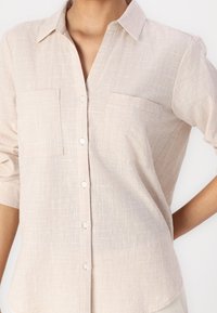 Chemise beige claire à boutons avec un motif à carreaux texturé, un col, et deux poches sur la poitrine. Fabriquée dans un tissu doux avec des manches courtes.