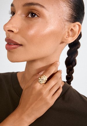 Close-up af en kvinde med flettet hår iført en mørk skjorte, der viser en stor, tekstureret guldring på hånden nær hendes hals.