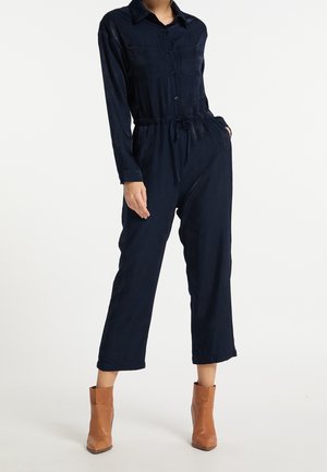 Kvinde iført en marineblå heldragt med lange ærmer og frontlommer samt tanfarvede ankelstøvler, stående mod en hvid baggrund.