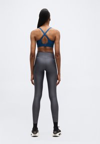 Γυναίκα που στέκεται πλάτη, φοράει ένα μπλε αθλητικό σουτιέν με σταυρωτές λωρίδες, γκρι εφαρμοστά κολάν, μαύρες κάλτσες και μαύρα-λευκά αθλητικά παπούτσια.