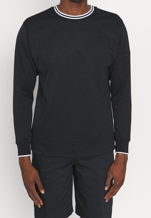 Mann trägt ein schwarzes Langarmshirt mit weißem Besatz an Kragen und Bündchen, kombiniert mit schwarzen Shorts vor einem einfarbigen Hintergrund.