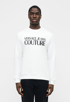 Homme portant un sweat-shirt blanc avec le texte "Versace Jeans Couture", debout devant un fond gris clair uni.