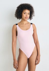 Maillot de bain une pièce de couleur rose clair avec un décolleté en U, des bretelles noires, des jambes échancrées et un tissu lisse et texturé.