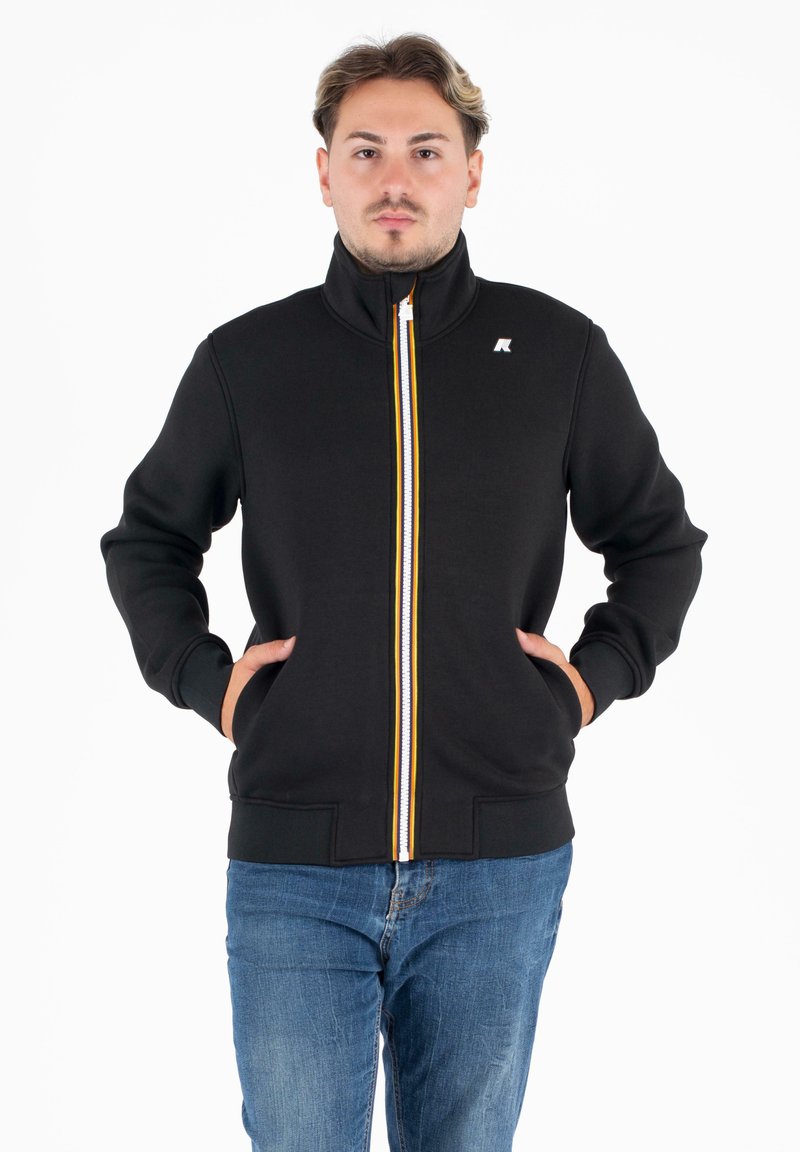 Zwarte zip-up jas met hoge kraag, voorzien van een witte en gele streep langs de rits. Zacht materiaal, zijzakken en ribgebreide boorden.