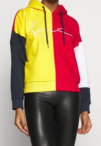 Donna che indossa un hoodie color block con sezioni gialle, rosse, bianche e blu navy, abbinato a leggings neri in ecopelle.