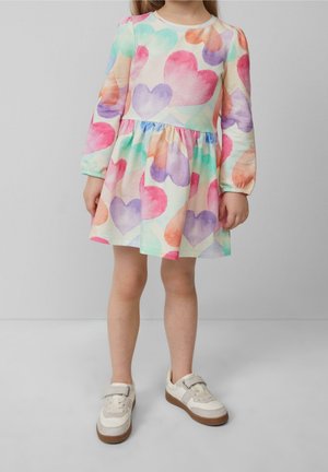 Kind draagt een lange mouw jurk met veelkleurige hartjespatronen en beige sneakers met bruine zolen, staand tegen een effen grijze achtergrond.