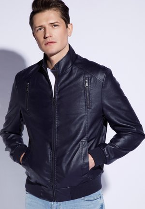 Marineblaue Lederbomberjacke mit Stehkragen, zwei gefütterten Brusttaschen mit Reißverschluss und gerippten Bündchen. Glatte Textur, taillierte Passform und minimalistischer Stil.