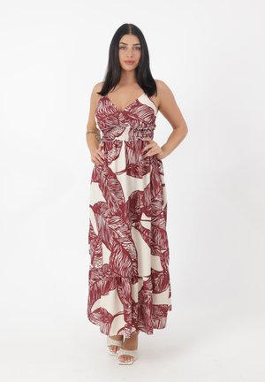 Mujer de pie con las manos en las caderas, vistiendo un vestido largo blanco sin mangas con patrones de hojas rojas y tacones abiertos blancos contra un fondo blanco.