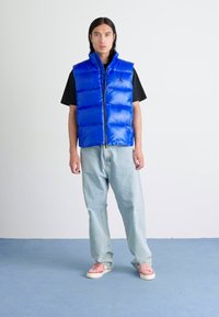 Gilet sans manches bleu matelassé avec une fermeture éclair à l'avant, porté par-dessus une chemise noire. Le modèle l'associe avec un jean bleu clair et des baskets roses.