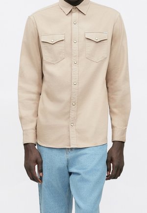 Chemise beige à boutons avec deux poches poitrine et pressions métalliques, portée sans être rentrée sur un jean bleu clair sur une personne à la peau foncée.
