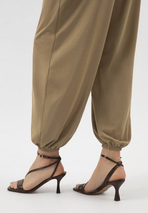 Füße, die braune Riemchen-High-Heel-Sandalen tragen, kombiniert mit weiten beigen Hosen, die an den Knöcheln gerafft sind.