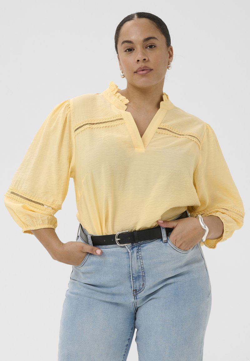Femme portant un chemisier jaune clair à volants avec des inserts en dentelle et un jean taille haute bleu clair, posant les mains dans les poches.