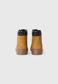 Botas de tobillo de ante marrón con un collar de cuero oscuro, suela de goma y detalle de costura blanca. El diseño presenta una textura suave y una estructura resistente.