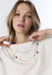 Felpa bianca con gioielli sovrapposti: una catena d'oro con ciondoli (lucchetto, cuore, chiave) e perline che formano la scritta "NO DRAMA."