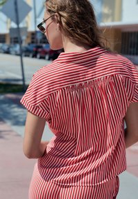 Piros-fehér csíkos rövid ujjú ing, hátul gyűrt részlettel és sima textúrával. Kerekített szegély, laza szabás.