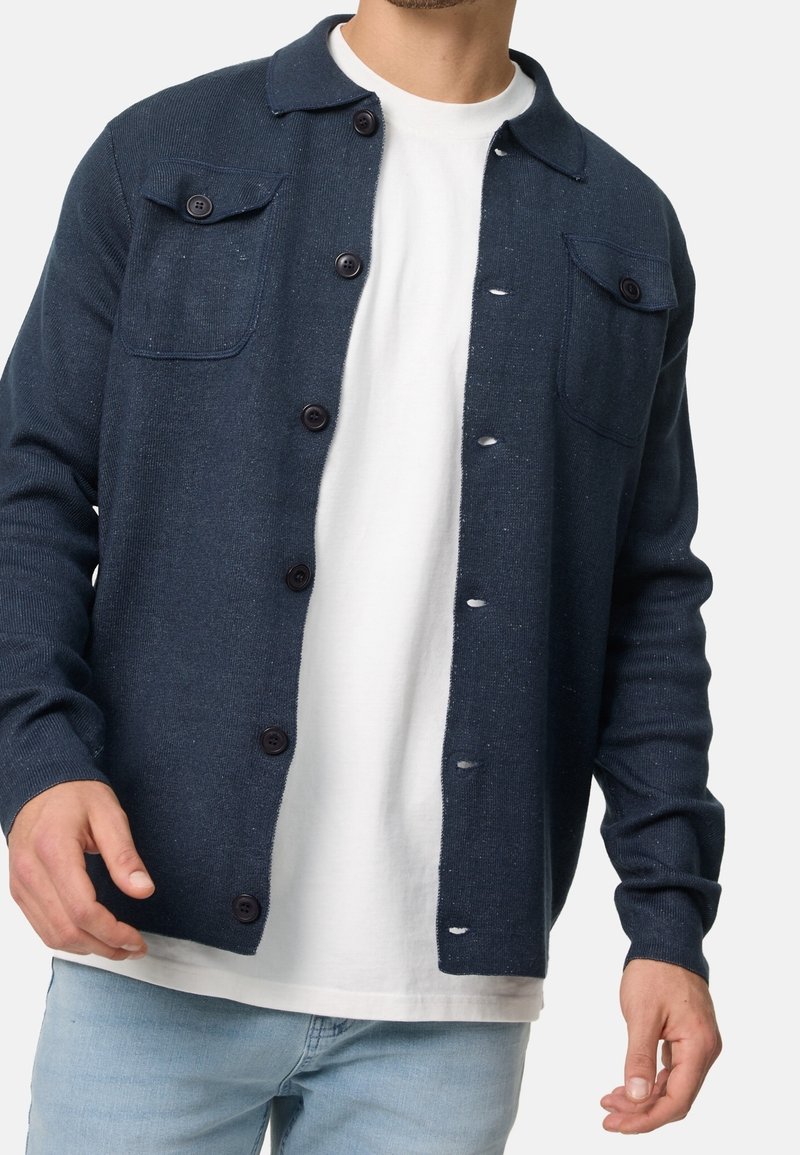 Homme portant un cardigan bleu foncé boutonné avec des poches poitrine par-dessus un t-shirt blanc et un jean bleu clair.
