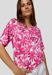 Pink floral short-sleeve top with a white background, featuring large stylized flowers and a textured fabric finish.