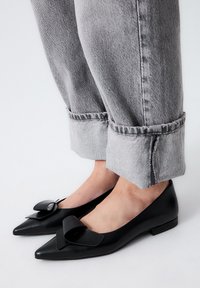 Zapatos planos de cuero negro con puntera afilada, adornados con un gran lazo decorativo en cada zapato. Combinados con unos pantalones vaqueros grises enrollados, que dejan ver los zapatos.